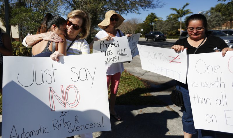 FILE- Cathy Kuhns, right, hugs Ana Paula Lopez, left, as they stand on a street corner holding up anti gun signs in Parkland, Fla., on Saturday, Feb. 17, 2018. As families begin burying their dead, authorities are questioning whether they could have prevented the attack at the high school where a gunman, Nikolas Cruz, took several lives. (AP Photo/Brynn Anderson)