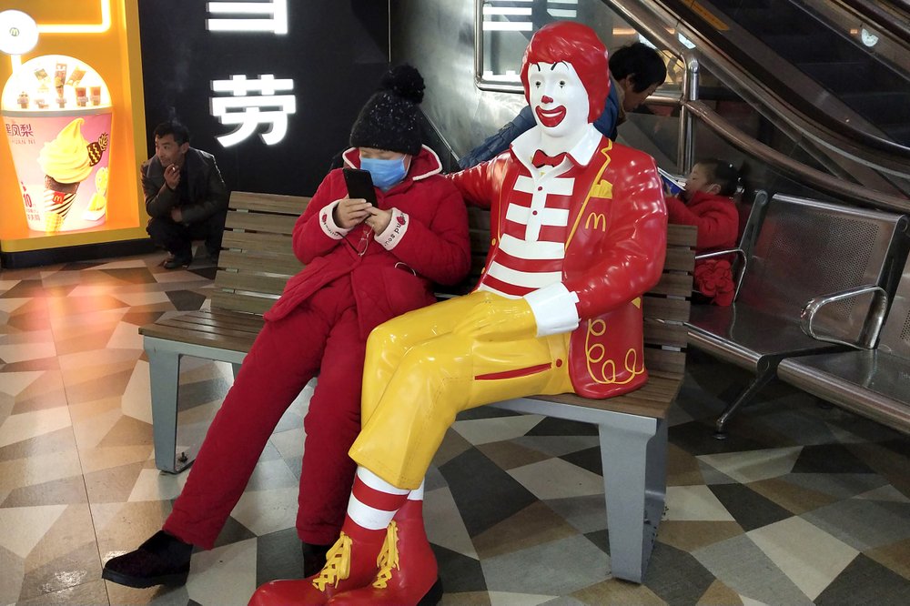 A woman wears a face mask and she uses her smartphone as she sits next to a statue of Ronald McDonald at a McDonald's restaurant in Suzhou in eastern China's Jiangsu Province, Sunday, Jan. 26, 2020. A new viral illness being watched with a wary eye around the globe accelerated its spread in China on Sunday with 56 deaths so far. (Chinatopix via AP)